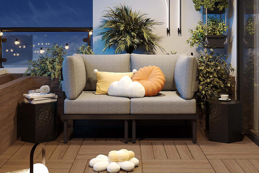 Gray sofa with colorful cushions on a wooden deck at night, surrounded by plants and lights.