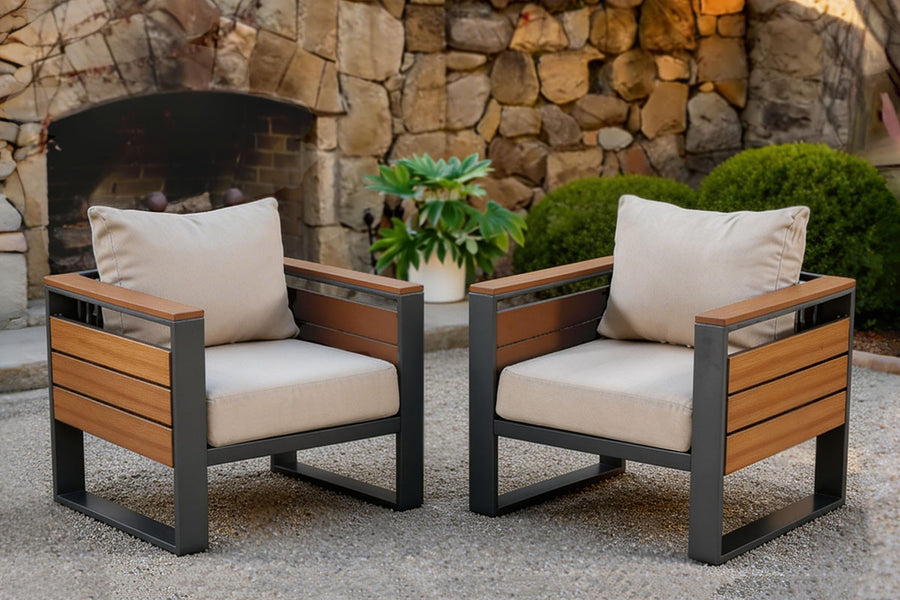 Two wooden finish outdoor chairs with beige cushions in front of a stone wall.