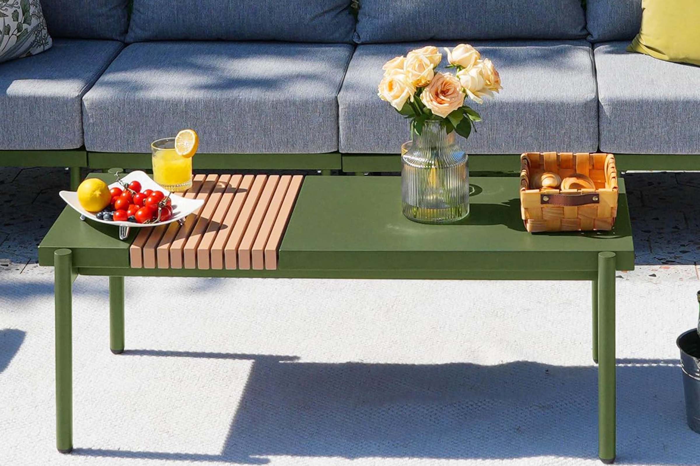 Green outdoor coffee table with a wooden top, fruit, flowers, and a basket on a patio.#color_green